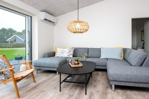 Moderne opholdsstue i 8 personers feriehus i Ansager, Danmark, med grå hjørnesofa og store vinduer.