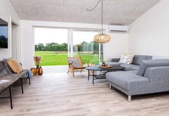 Modernes Wohnzimmer in einem Ferienhaus in Ansager, Dänemark, mit grauem Sofa, großen Fenstern und Blick ins Grüne.