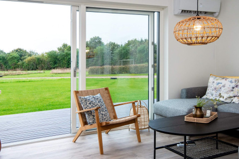 Moderne zithoek in vakantiehuis met schuifdeuren, uitzicht op groen gazon, grijze bank en houten stoel.