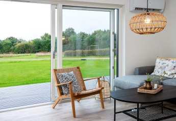 Moderne zithoek in vakantiehuis met schuifdeuren, uitzicht op groen gazon, grijze bank en houten stoel.