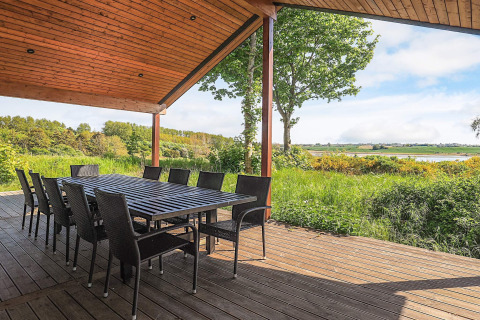 Overdekt houten terras met ruime eettafel en stoelen, uitkijkend op groen en meer in Millinge, Denemarken.