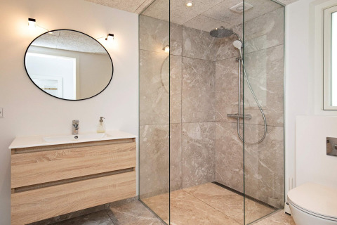 Moderne badkamer met ruime inloopdouche, rond spiegel en houten meubel in een vakantiewoning in Millinge.