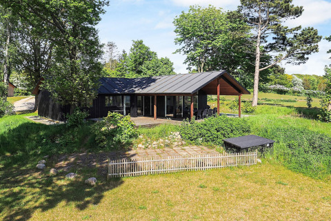 Moderne feriebolig i Millinge, Danmark, omgivet af grønne træer og have med overdækket terrasse.