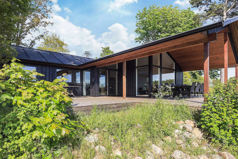 Moderne 12-personers feriehus i Millinge, Danmark, med stor terrasse, sort facade og grønne omgivelser.