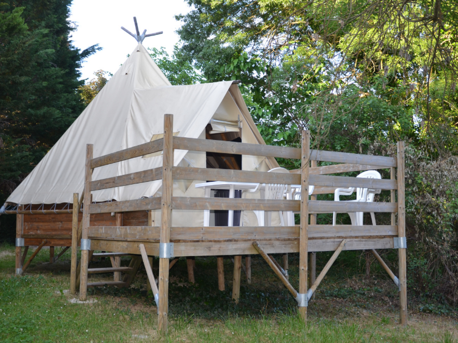 Een Tipi Tent met houten terras en witte stoelen, omgeven door bomen, gefotografeerd op een zonnige dag.