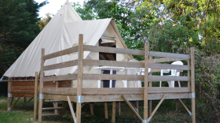 Un tipi nommé Tipi Tent avec terrasse en bois et chaises blanches, entouré d’arbres lors d’une journée claire.