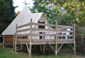 Een Tipi Tent met houten veranda en witte stoelen, omgeven door groene bomen, genomen bij daglicht.