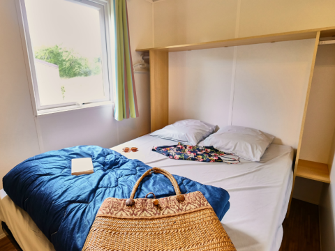 Dormitorio en Bungalowtent de Ludo Camping, Francia, con cama doble, ventana y bolso de mimbre.