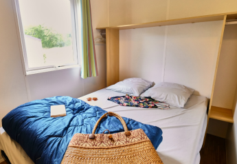 Dormitorio en Bungalowtent de Ludo Camping, Francia, con cama doble, ventana y bolso de mimbre.