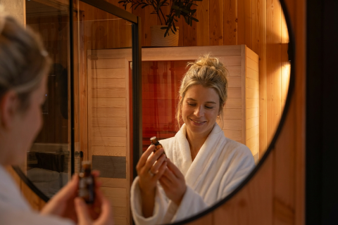Woman in a white bathrobe looks at a bottle of oil in front of a mirror at Wellness Duynzicht, Sandberghe.