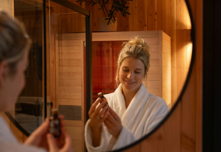 Woman in a white bathrobe looks at a bottle of oil in front of a mirror at Wellness Duynzicht, Sandberghe.