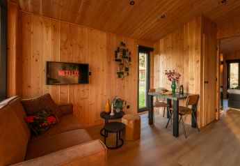 Gezellig houten lodge-interieur met sofa, tv met Netflix, eettafel voor twee en tuinzicht.