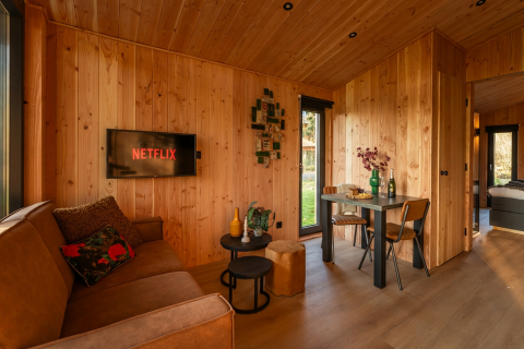 Interno di lodge accogliente in legno con divano, TV su Netflix, tavolo da pranzo e vista sul giardino.