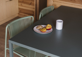 Rincón de comedor en casa pequeña con mesa negra, silla de alambre, plato de galletas, fruta y taza blanca.