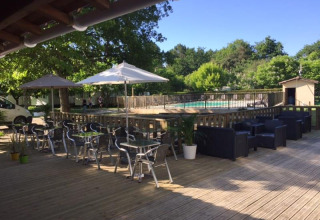 Terrazza all'aperto con tavoli, sedie e ombrelloni accanto a una piscina recintata in un ambiente verde.