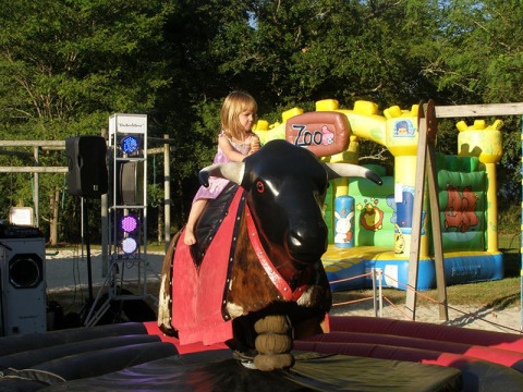 A young girl rides a mechanical bull at Campsite Les Franquettes with a colorful bouncy castle behind her.