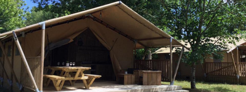 Safari-Zelt Lodge Woody 27 mit überdachter Terrasse, Holztisch und grünem Garten im Hintergrund.