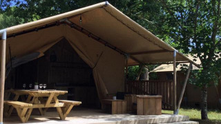 Safari-Zelt Lodge Woody 27 mit überdachter Terrasse, Holztisch und grünem Garten im Hintergrund.