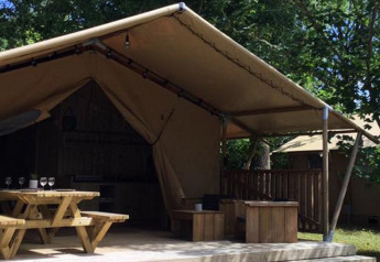 Tienda safari Lodge Woody 27 con terraza de madera cubierta y bancos al aire libre entre árboles.