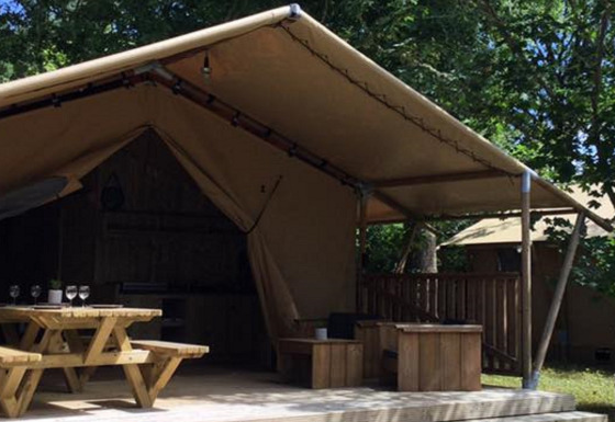 Safari-Zelt Lodge Woody 27 mit überdachter Terrasse, Holztisch und grünem Garten im Hintergrund.