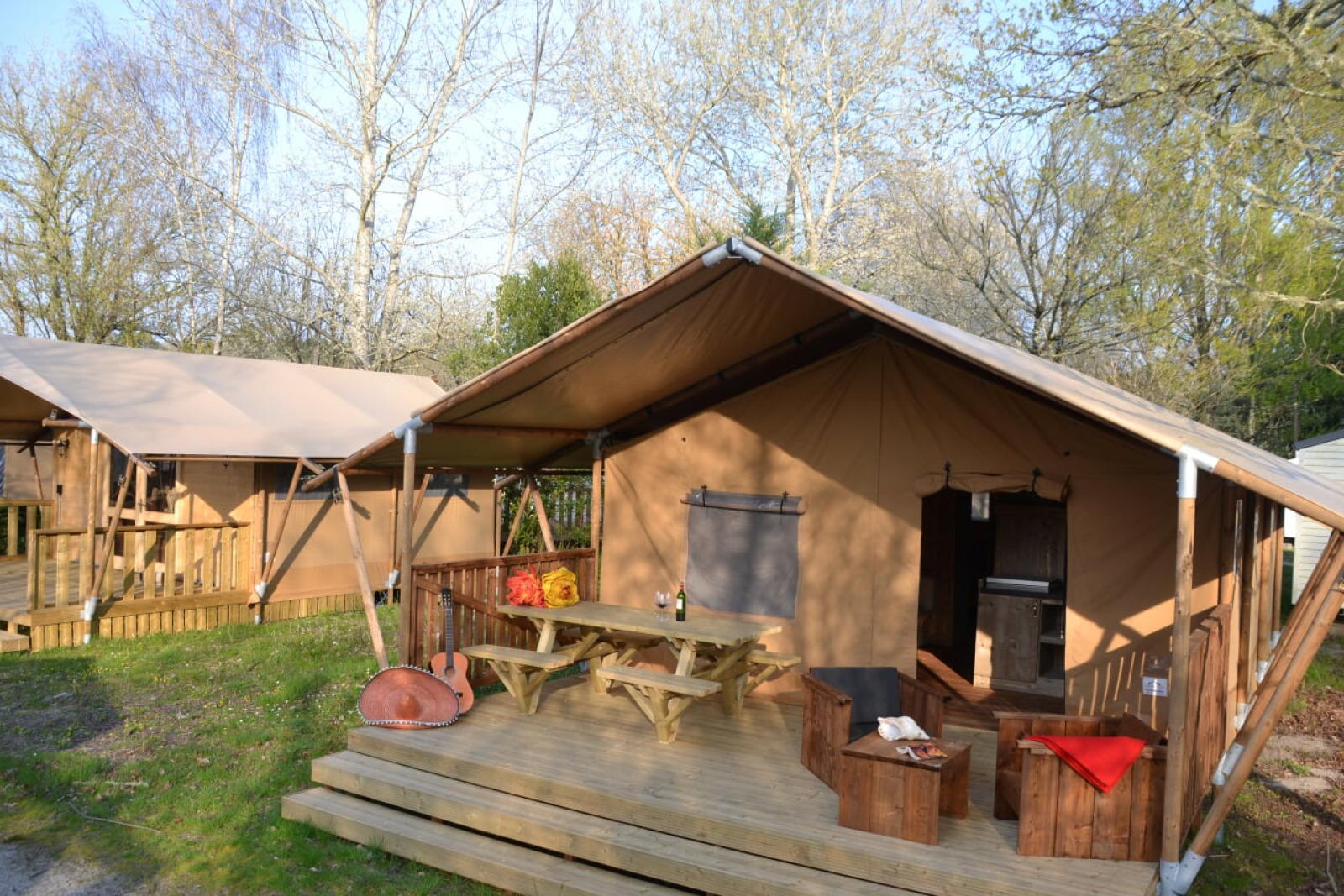 Safaritent Lodge Woody 27 met houten veranda, picknicktafel, gitaar en zitplaatsen te midden van bomen.