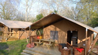 Safari tent Lodge Woody 27 with wooden deck, picnic table, guitar, and rustic seating amid trees.
