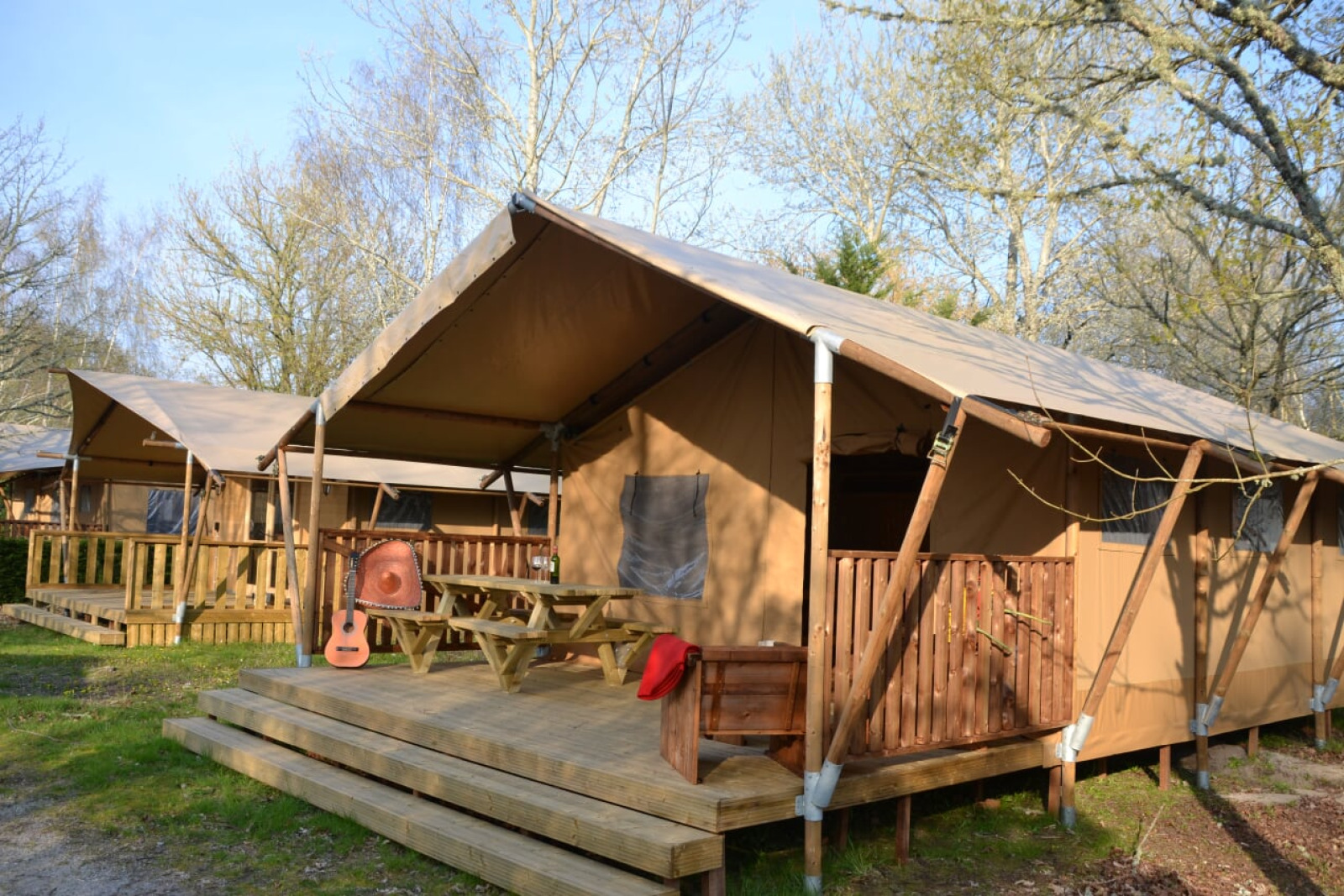 Safari-tent Lodge Woody 38 met houten terras, picknicktafel, gitaar en omgeven door bomen.
