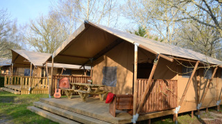 Tenda safari Lodge Woody 38 con terrazza in legno, tavolo da picnic, chitarra e circondata da alberi.