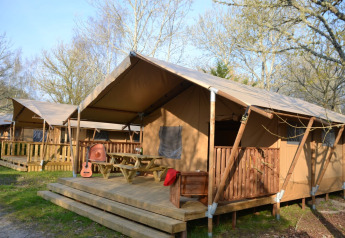 Safari-Zelt Lodge Woody 38 mit Holzterrasse, Picknickbank, Gitarre und umgeben von Bäumen im Freien.