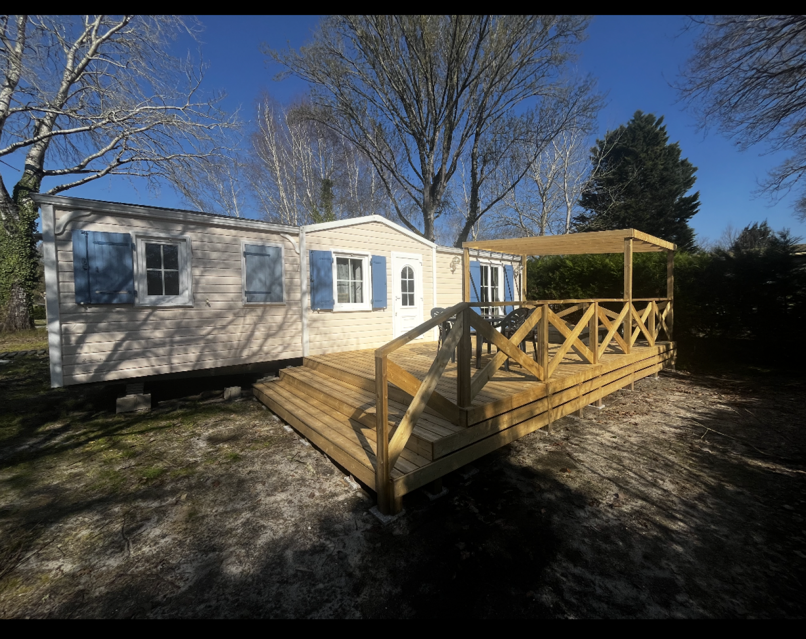 Premium Mobile Home Spa met houten terras, blauwe luiken en bomen eromheen onder een heldere lucht.