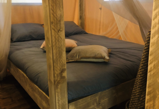 Bett mit Holzrahmen und Moskitonetz in einem Safari-Zelt Lodge Woody 27 auf dem Campingplatz Les Franquettes.