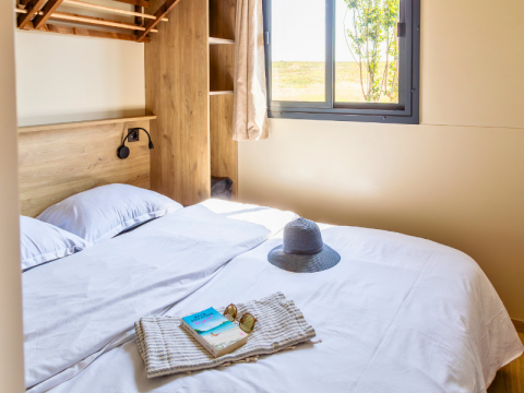 Lichte slaapkamer in een Premium chalet op Camping Les Tamaris in Frankrijk, met hoed en boek op bed.