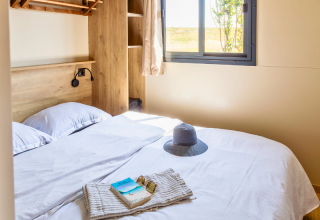 Lichte slaapkamer in een Premium chalet op Camping Les Tamaris in Frankrijk, met hoed en boek op bed.