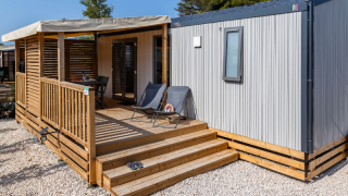 Premium mobile home with wooden deck, sun loungers, and garden furniture, surrounded by gravel and trees
