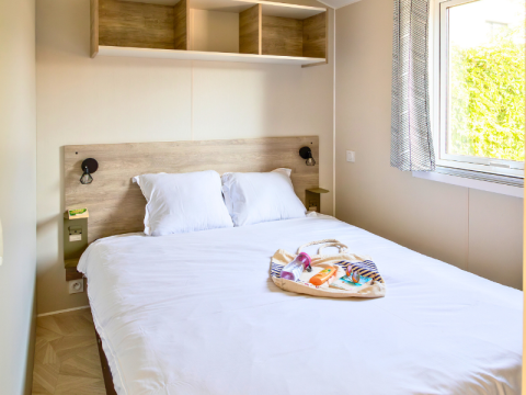 Premium mobile home bedroom with double bed, window, and bag set at Campsite Les Tamaris in France.