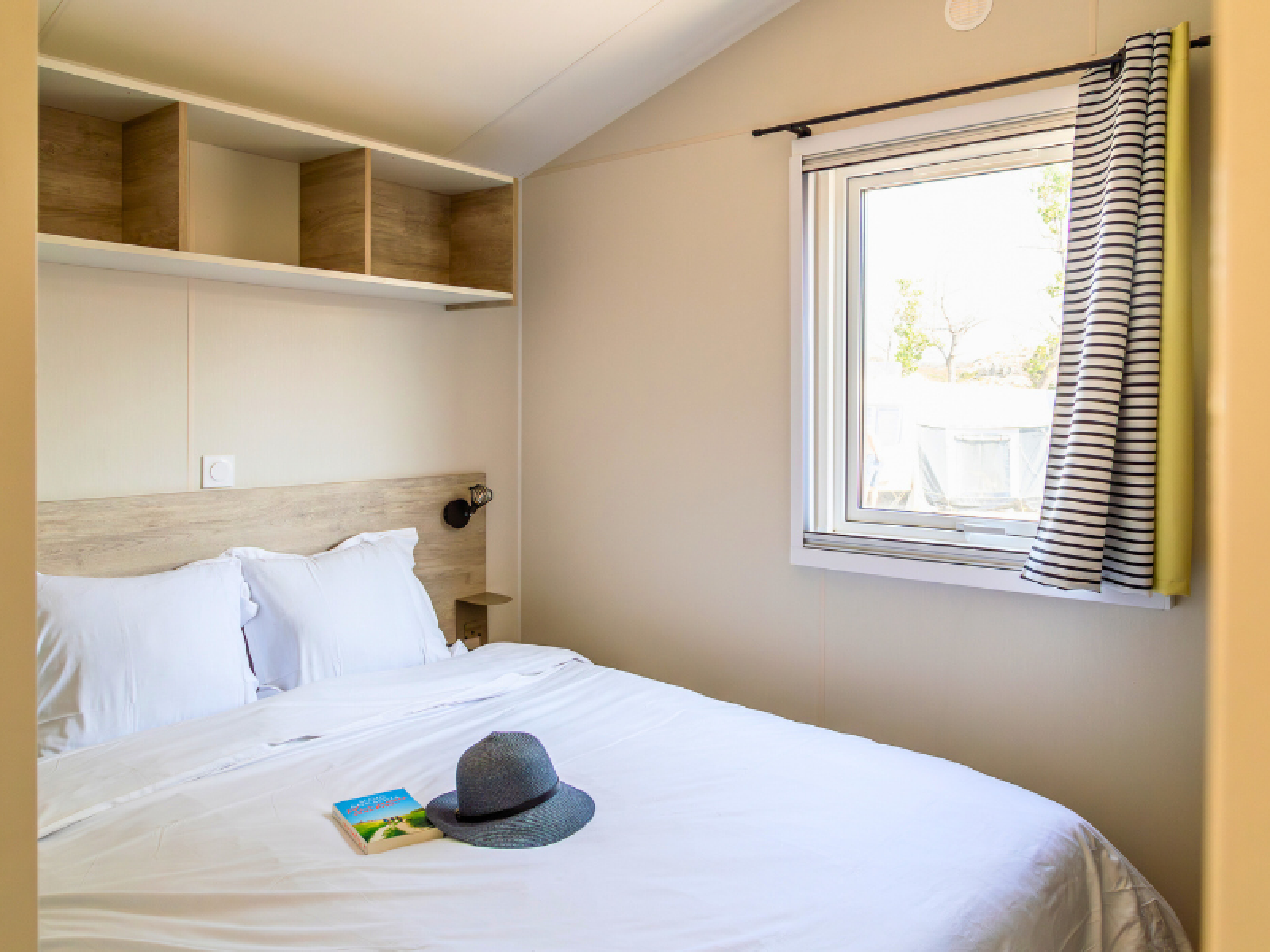 Slaapkamer in Premium Mobile Home 'Large' op Camping Les Tamaris, Frankrijk, met bed, raam en boek.