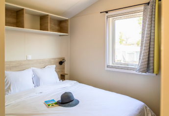 Dormitorio en Premium Mobile Home «Large» en el camping Les Tamaris, Francia, con cama, ventana y libro.