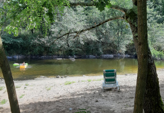 Peaceful riverside beach with a green chair, trees, and a person canoeing, Ushuaïa Villages Le Viaduc, France.