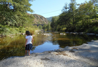 A child stands by a clear river near Ushuaïa Villages Le Viaduc, with a viaduct and hills in the background.