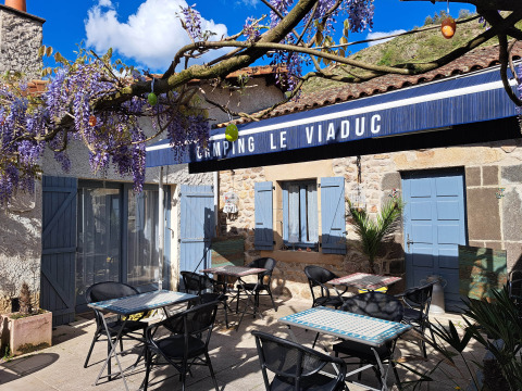 Area esterna del Camping Le Viaduc con tavoli, sedie, persiane blu e fiori di glicine viola in Francia.