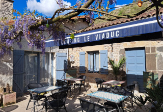 Area esterna del Camping Le Viaduc con tavoli, sedie, persiane blu e fiori di glicine viola in Francia.