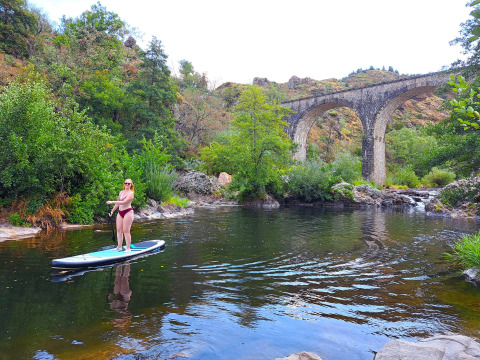Woman paddleboarding on a river near a historic stone viaduct at Ushuaïa Villages Le Viaduc campsite, France.