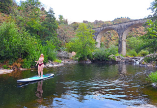 Woman paddleboarding on a river near a historic stone viaduct at Ushuaïa Villages Le Viaduc campsite, France.
