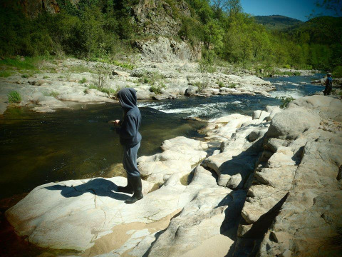 A person in a hoodie stands on rocks fishing by a scenic river at Ushuaïa Villages Le Viaduc campsite in France.