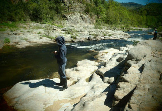 A person in a hoodie stands on rocks fishing by a scenic river at Ushuaïa Villages Le Viaduc campsite in France.