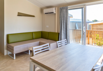 Moderne chaletstue med grøn sofa, spisebord, stole og adgang til terrasse på Camping Les Tamaris, Frankrig.