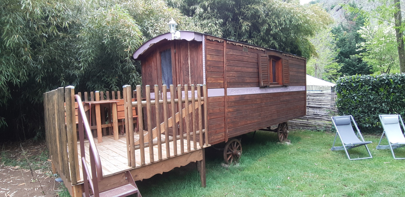 Tiny Wagon-hut bij Campsite Ushuaïa Villages Le Viaduc in Frankrijk met houten terras in een groene omgeving.