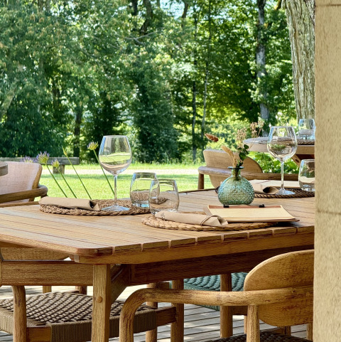 Outdoor wooden dining table set at La Nouvelle Croze campsite, France, with glasses and lush green scenery.