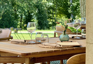 Outdoor wooden dining table set at La Nouvelle Croze campsite, France, with glasses and lush green scenery.