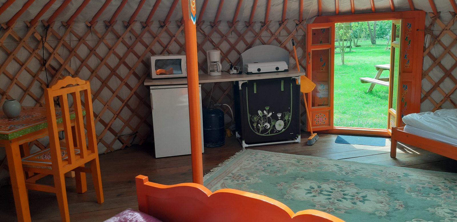 Interieur van een yurt met kleurrijk meubilair, keukentje en open deur met zicht op het groene gras.